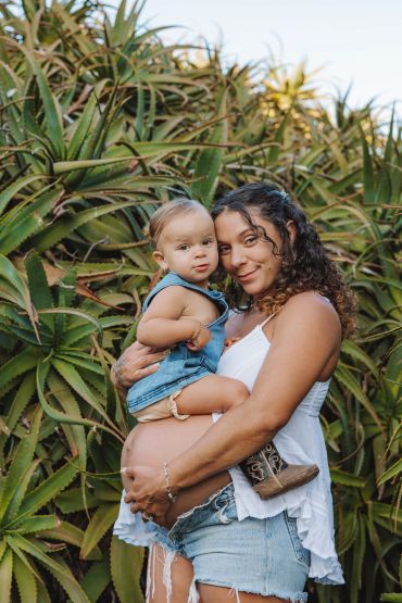 A pregnant woman holding a baby in front of aloe vera plants.