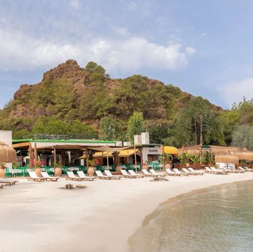 A tranquil beach resort with lounge chairs and a rocky hill backdrop under a partly cloudy sky.