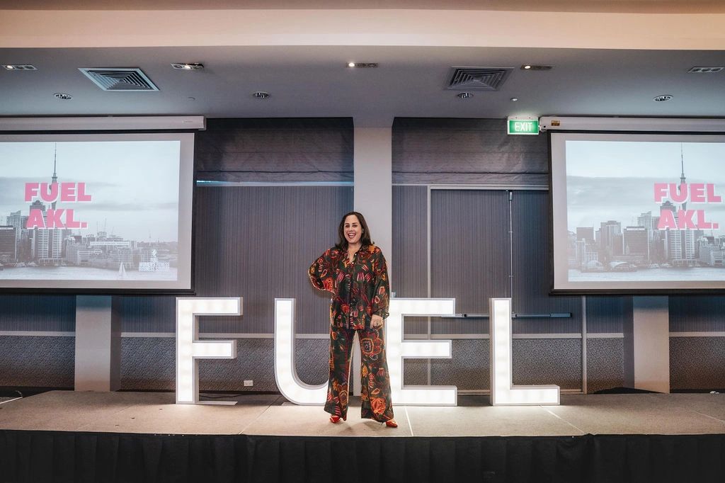 A woman stands confidently on stage with illuminated letters spelling FUEL.