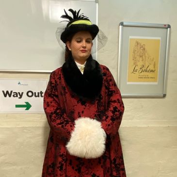 Kirsten is wearing a red and black dress with a white fluffy muff and a black and green hat.