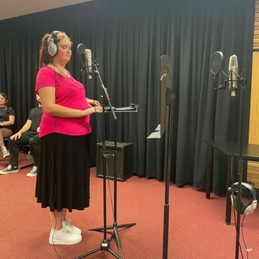 Kirsten is inside a recording studio in front of a microphone, wearing a black skirt and pink shirt.
