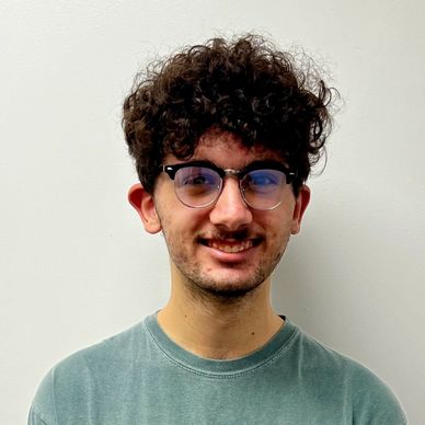 Young man with curly hair and glasses smiling against a plain wall.