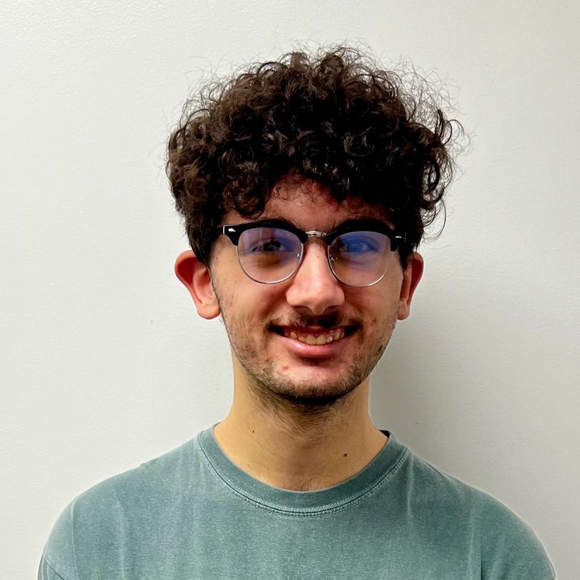 Young man with curly hair and glasses smiling against a plain wall.