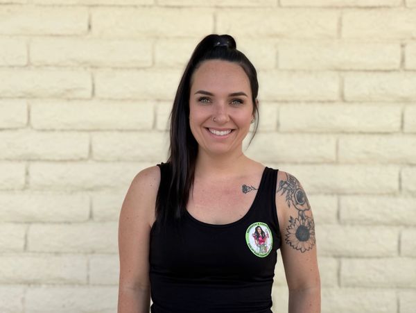 Smiling woman with tattoos in black tank top stands against a beige brick wall.
