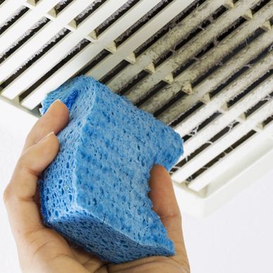 Hand holding a blue sponge near a dirty air vent.