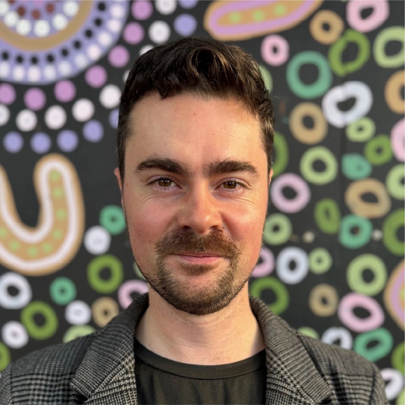 Man with a beard and mustache smiling in front of a colorful abstract background.