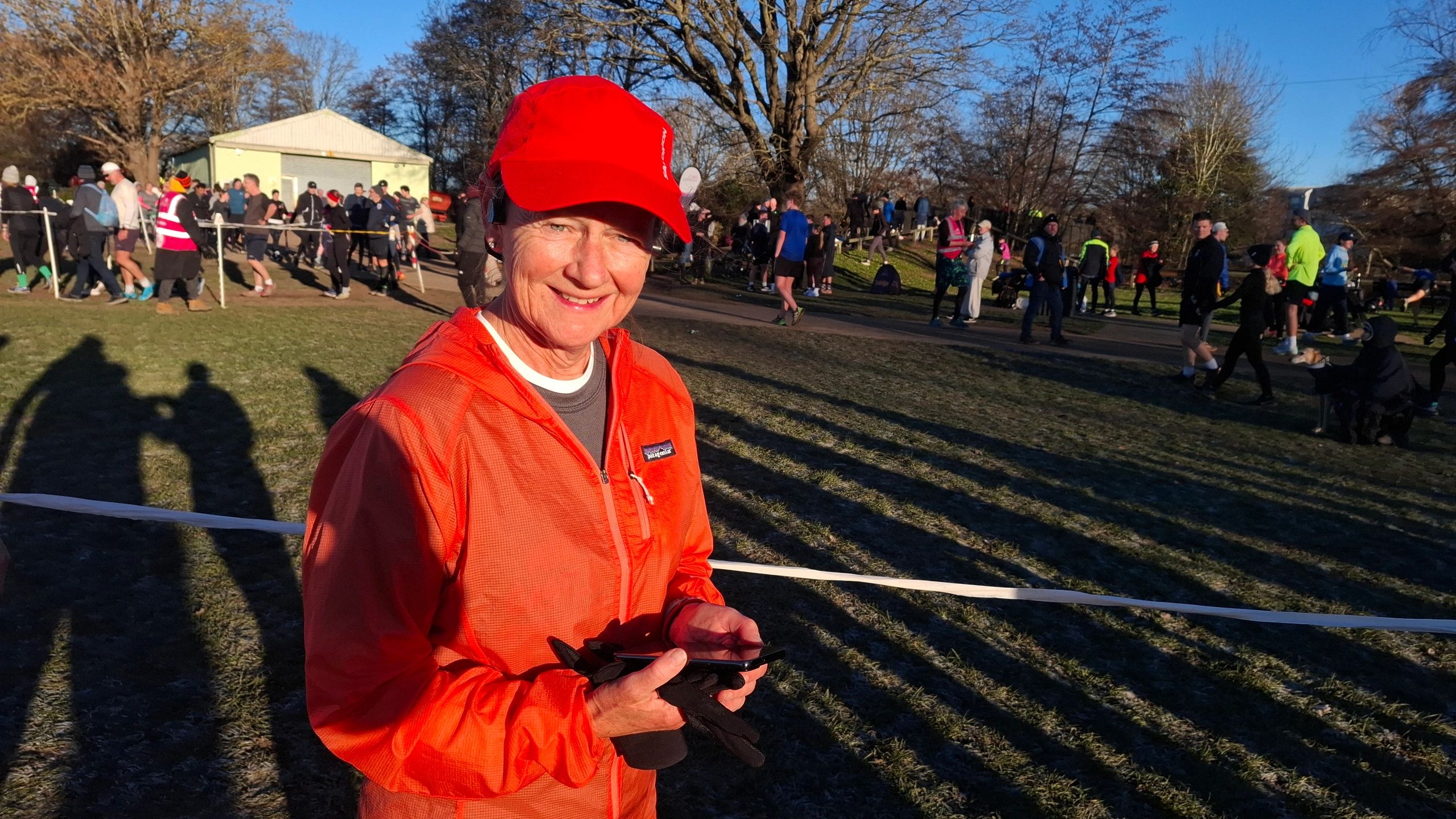 Woman in red jacket and cap smiling outdoors at a running event.