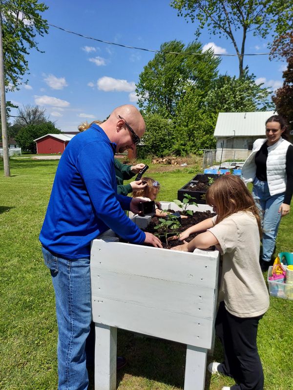 Adults and children planting seedlings in raised garden beds on a sunny day.