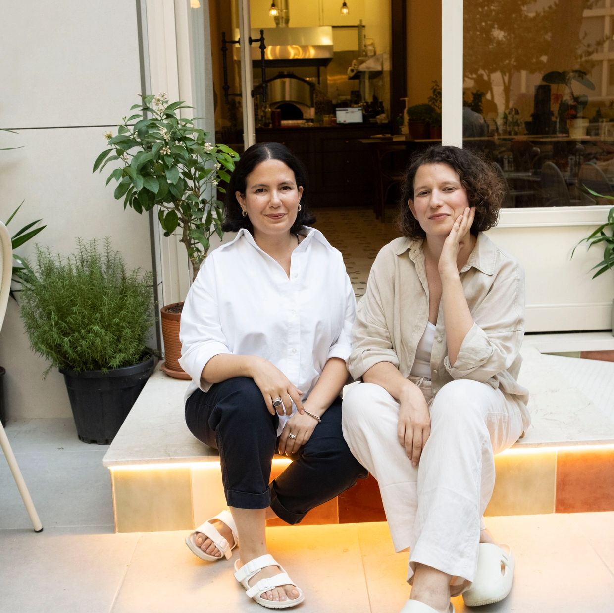 Two women sitting on a lit step outside a cozy café or restaurant.