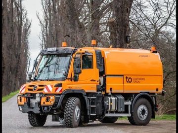 Orange utility truck on a cobblestone road near leafless trees.