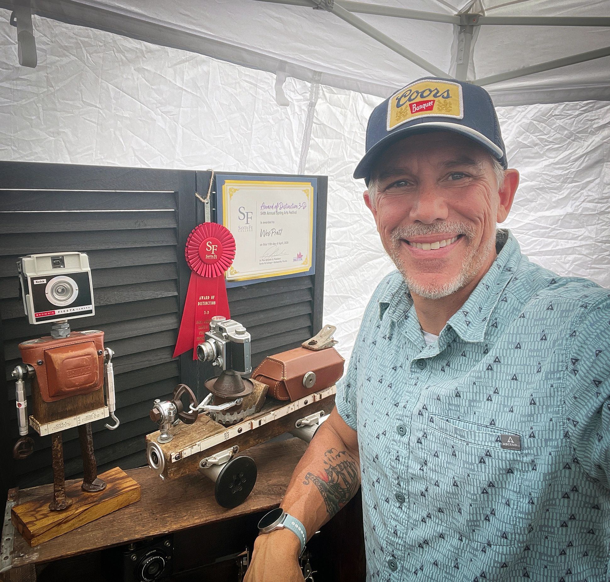 Man smiling beside vintage cameras and an award ribbon at an arts festival booth.