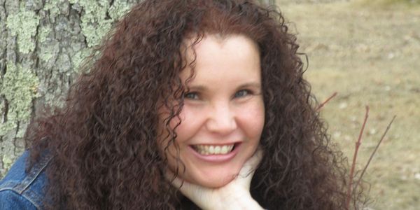 Smiling woman with curly hair leaning against a tree outdoors.