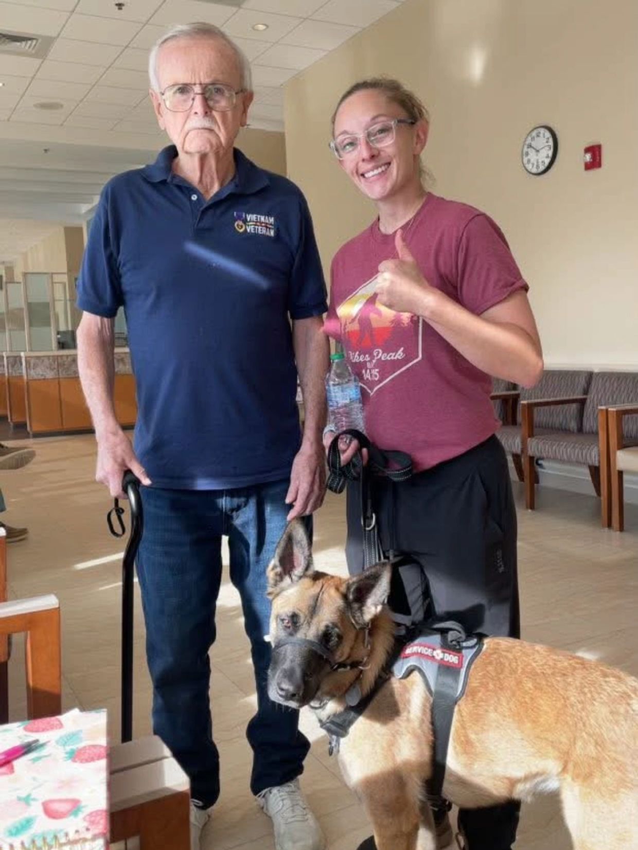 Older man with a cane stands beside a smiling woman and a service dog indoors.
