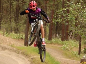 A child wearing a red helmet performs a bike jump in a forest trail.