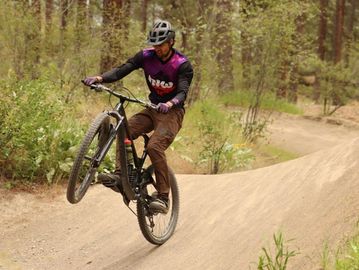 A cyclist performs a wheelie on a dirt trail in a forest.