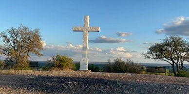 A large white cross on a hill with benches and trees under a blue sky.
