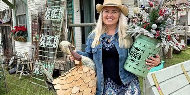 Smiling woman in cowboy boots holding decorative items in front of a rustic shop.