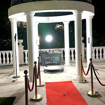 Illuminated photo booth setup with red carpet and prop table under white columns at night.