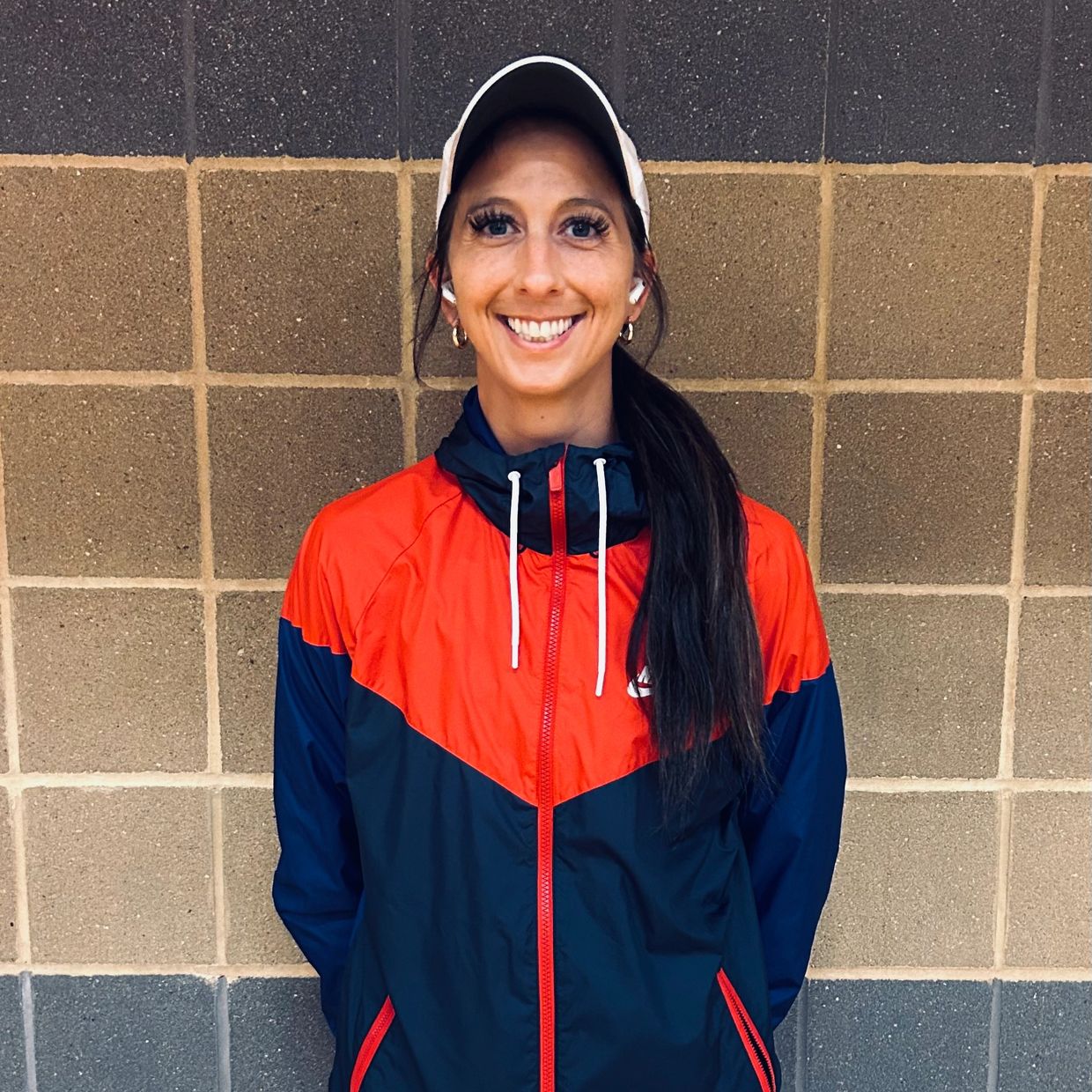 Smiling woman in a red and blue jacket and white cap against a tiled wall.