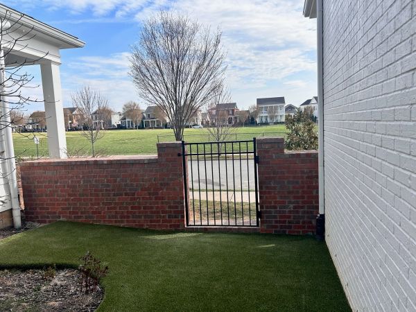 Brick wall with a metal gate overlooking a grassy field and houses.