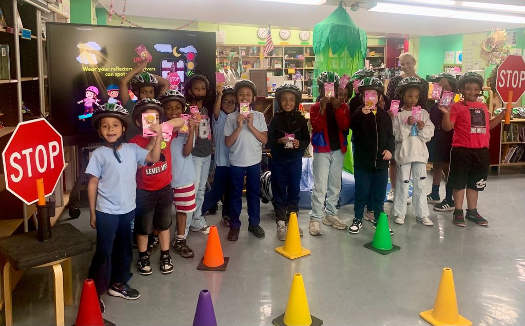 Children with helmets holding reflective gear in classroom.