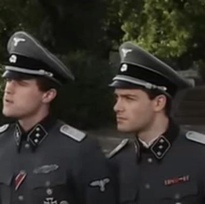 Two men in dark military uniforms with peaked caps looking to the side.