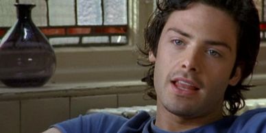 A man with dark hair and blue eyes sitting indoors, speaking.