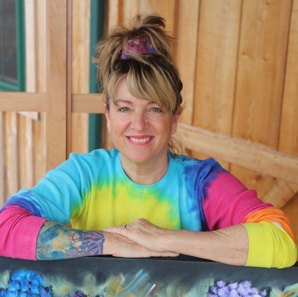 Smiling woman in a colorful tie-dye shirt with a tattoo on her arm. Artist Lisa fasulo