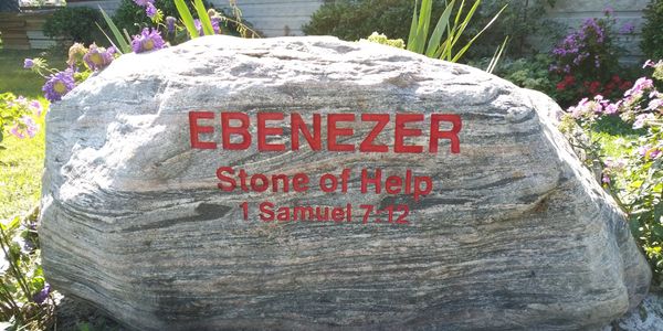 Large stone engraved with 'EBENEZER Stone of Help 1 Samuel 7:12' in a garden.