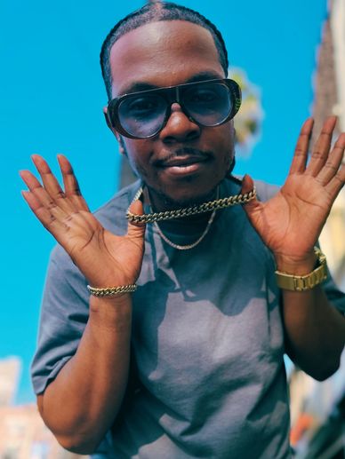A man wearing sunglasses and jewelry poses with his hands up.