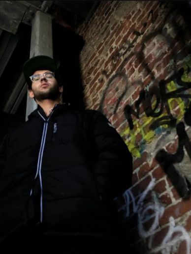 Young man in a black jacket stands against a graffiti-covered brick wall.
