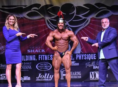 Bodybuilder posing in a Roman helmet on stage with two people pointing at him.