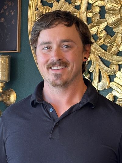 Smiling man with short hair and a black shirt standing indoors with ornate gold wall decor.