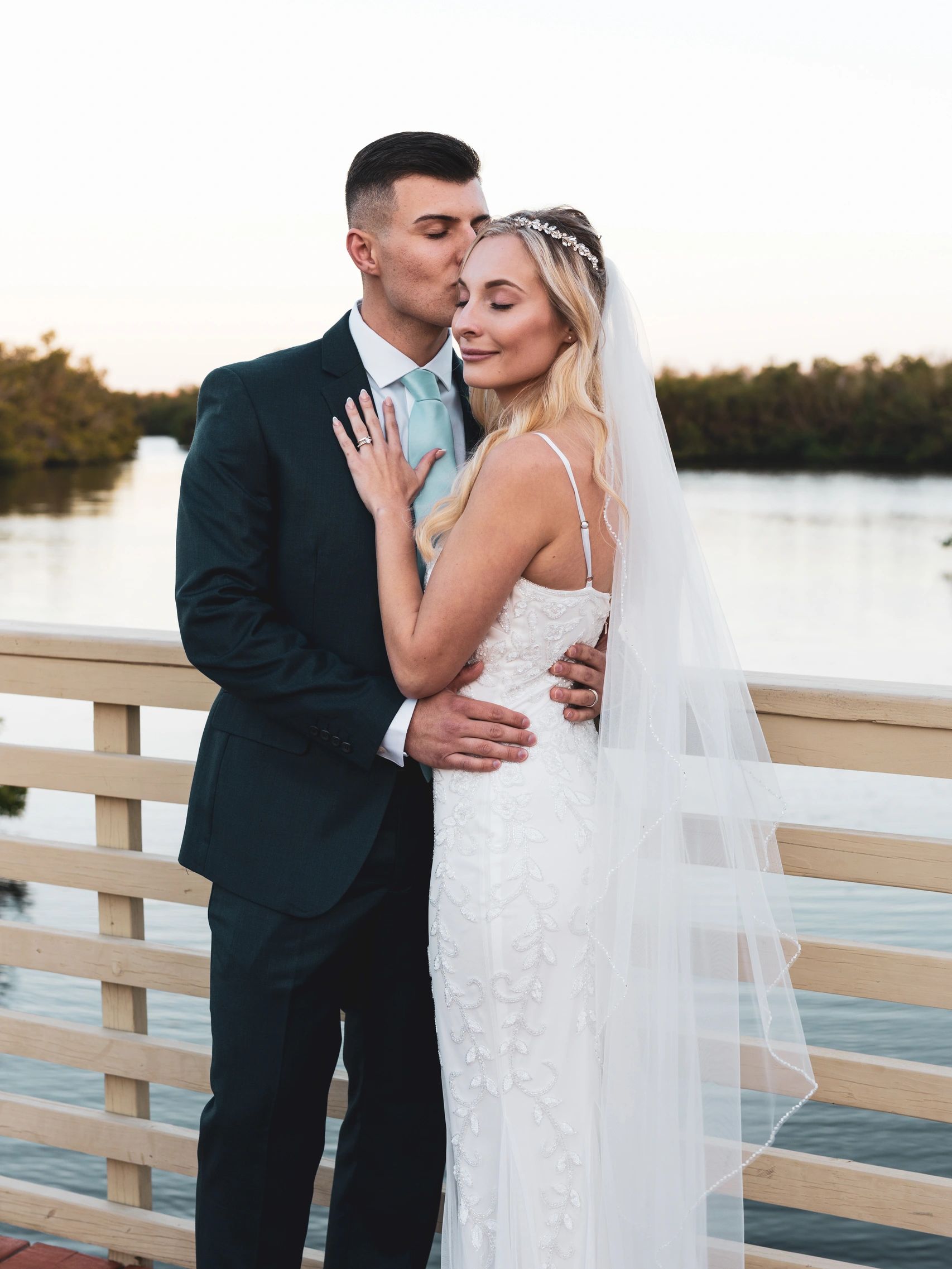 A groom kisses his bride on a bridge next to a lake.
