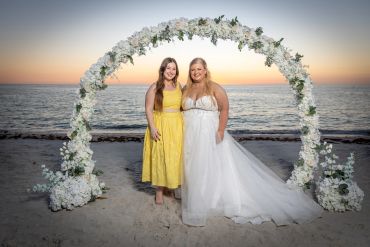 Two women pose under a floral arch on the beach at sunset, one in a wedding dress.
