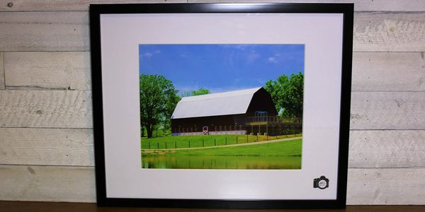 Framed photo of a barn with green trees and blue sky.