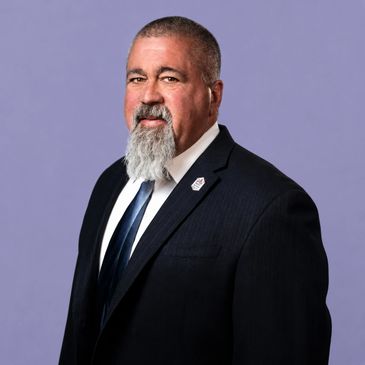 Portrait of a man in a suit with a gray beard against a purple background.