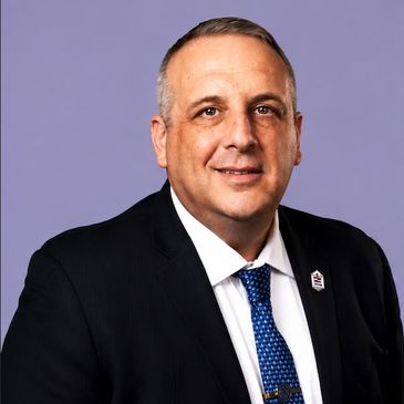 Professional portrait of a middle-aged man in a suit with a blue tie against a purple background.