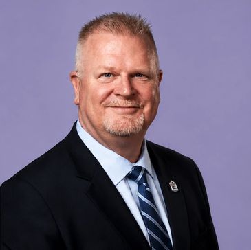 Professional headshot of a middle-aged man in a suit and tie against a purple background.