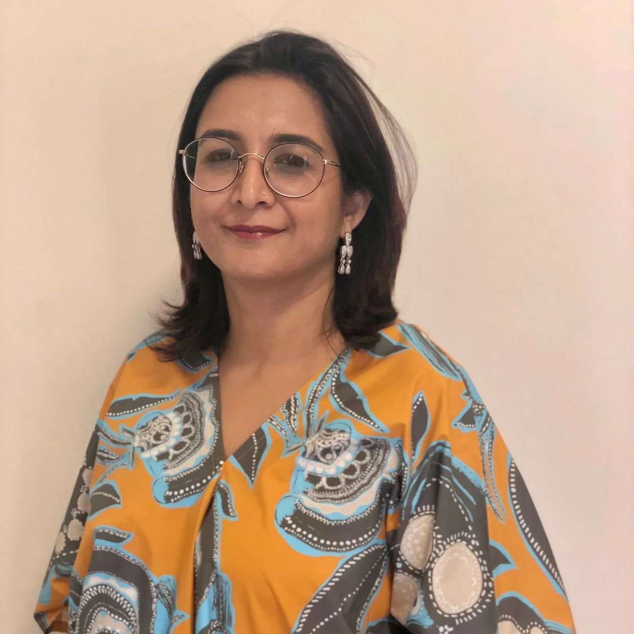 Woman with glasses wearing a colorful patterned top smiles gently.