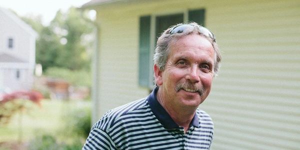 A smiling man in a striped shirt outdoors near a house.