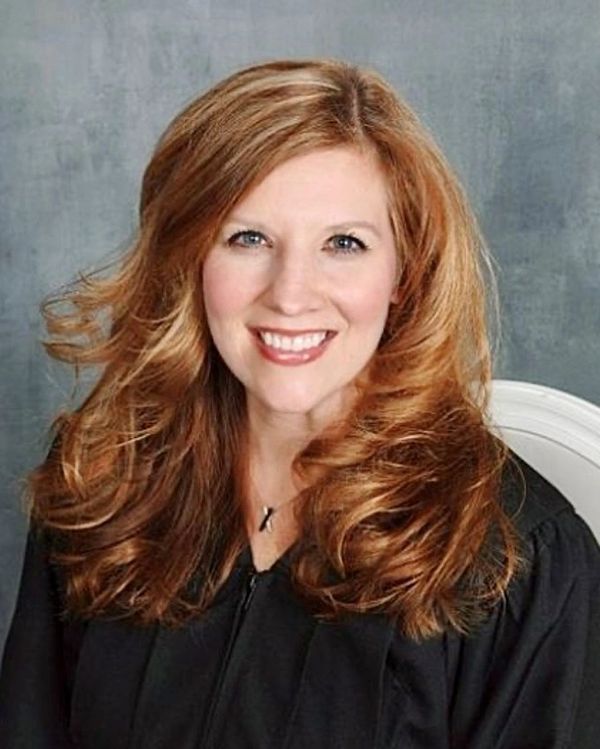 Smiling woman with wavy red hair in a black robe against a gray backdrop.