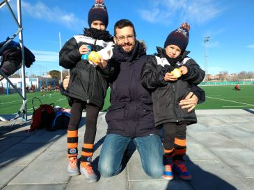 A man kneels with two children in sportswear and beanies on a sunny day.