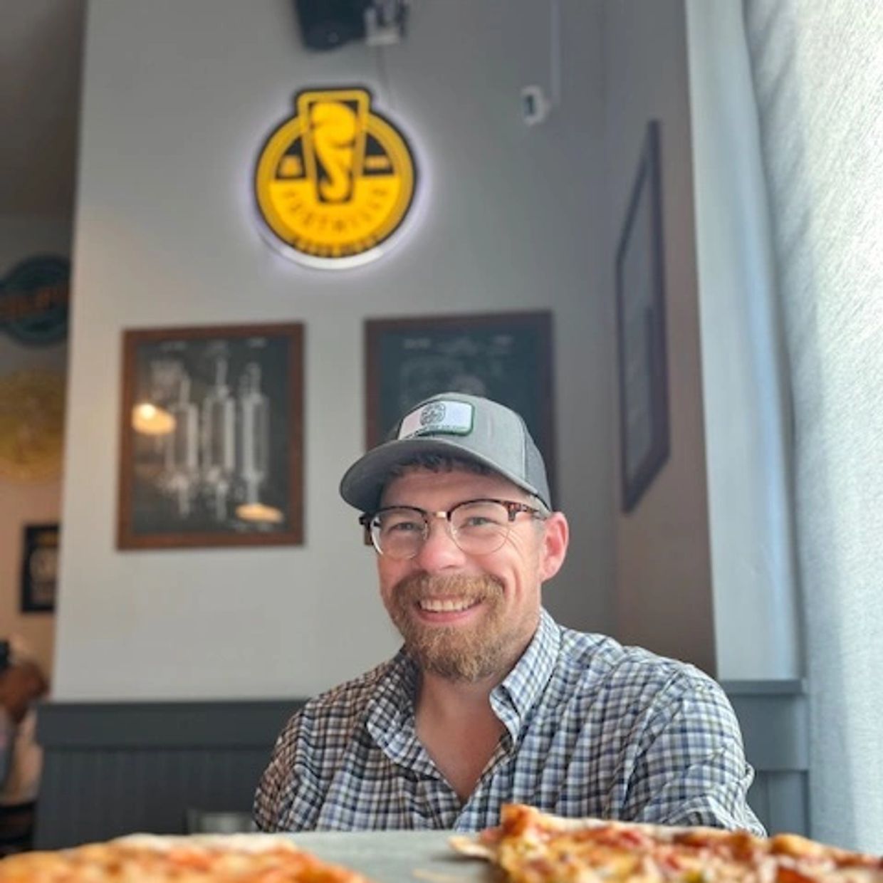 Smiling man with glasses and a cap sitting at a table with pizza.