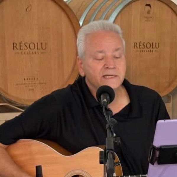 Man playing guitar and singing in front of wine barrels.