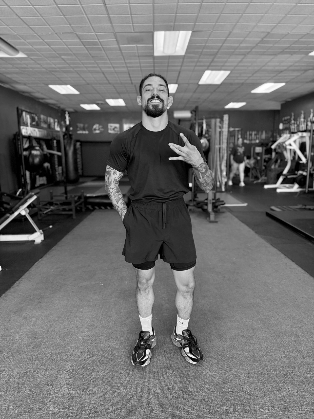Man in black athletic wear posing confidently in a gym with tattoos on his arms.