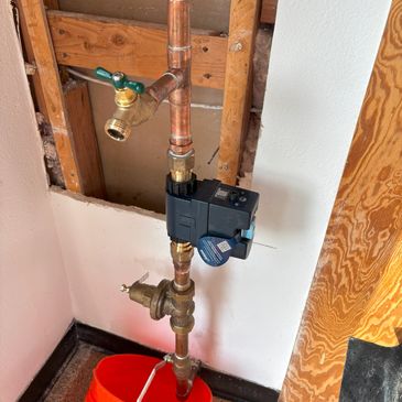 Copper piping with a valve and a black device above an orange bucket inside a wall cavity.