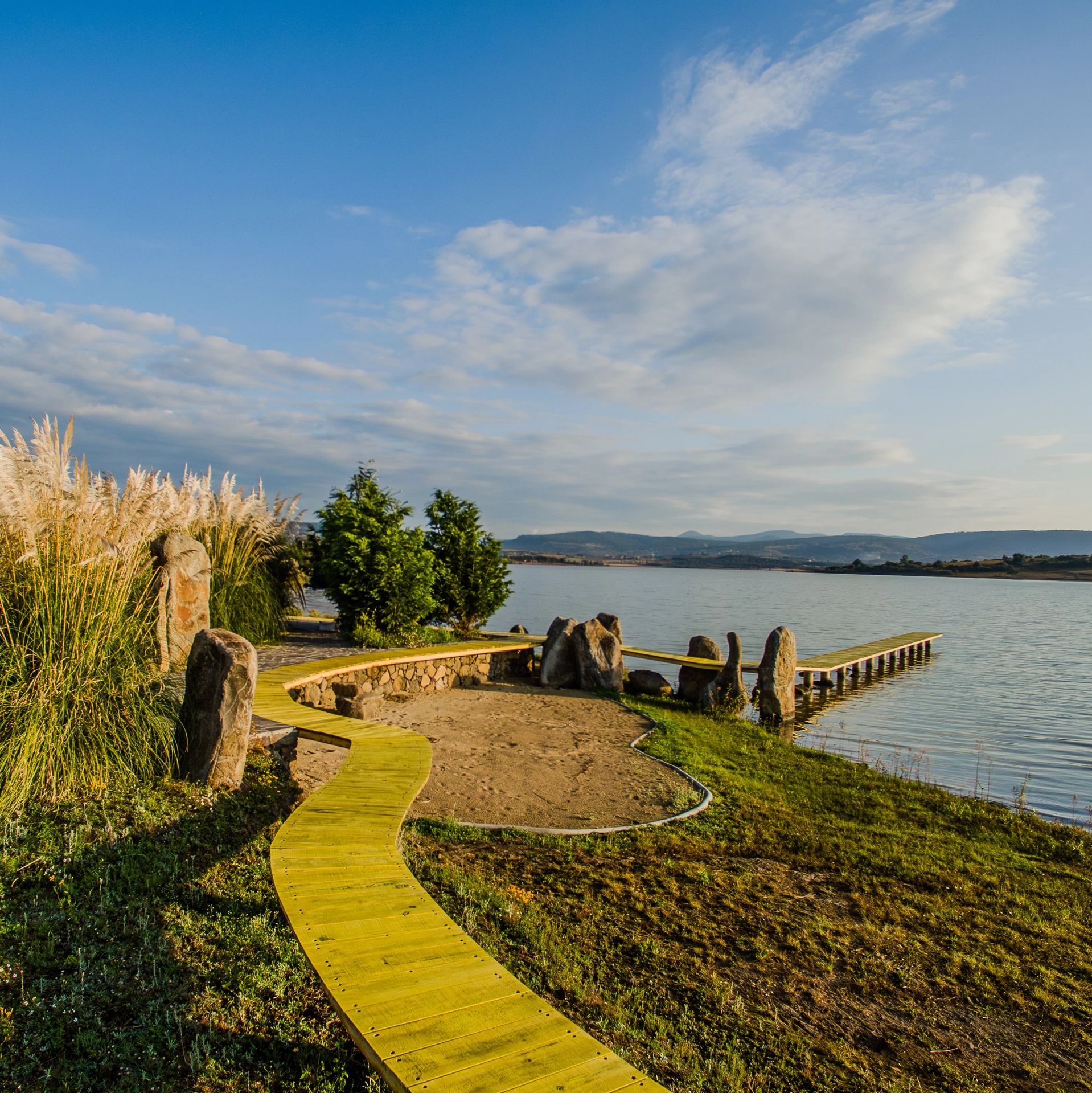 Yellow wooden pathway beside a lake with trees and stones under a blue sky.