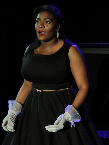 Iyana Colby standing mouth agape singing in a black 50's dress with gold accents and white gloves