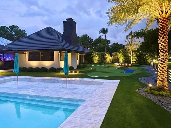 A modern backyard with a pool, palm tree, and putting green at dusk.